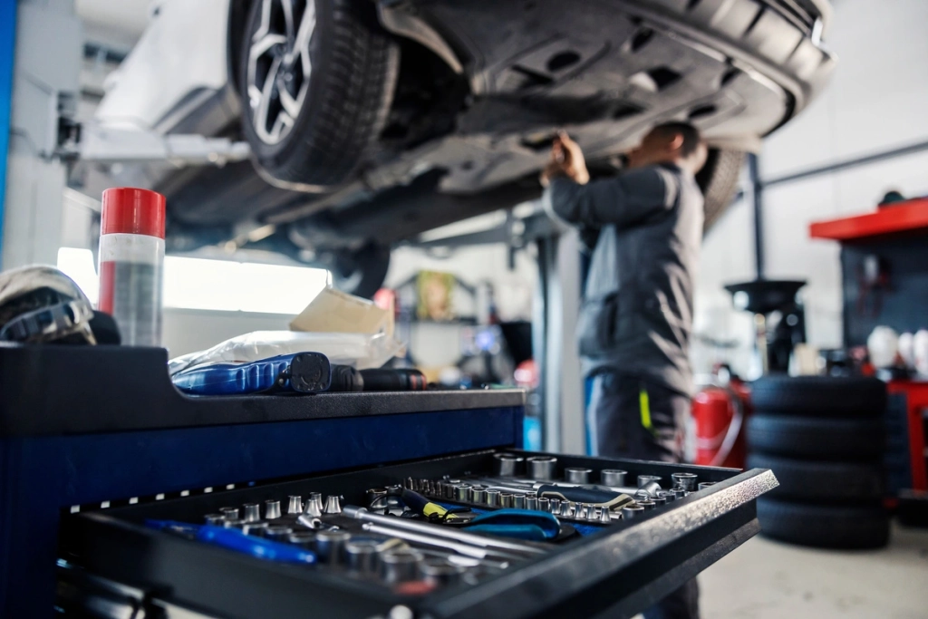 Intérieur d'un garage automobile avec un mécanicien travaillant sous une voiture surélevée, et au premier plan une boîte à outils ouverte avec divers outils visibles