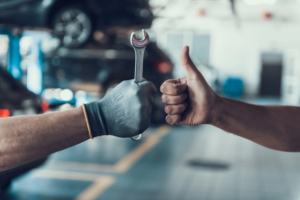 Gros plan sur deux mains dans un garage automobile : une main gantée tenant une clé à molette et l'autre main faisant un pouce en l'air, symbolisant un travail bien fait